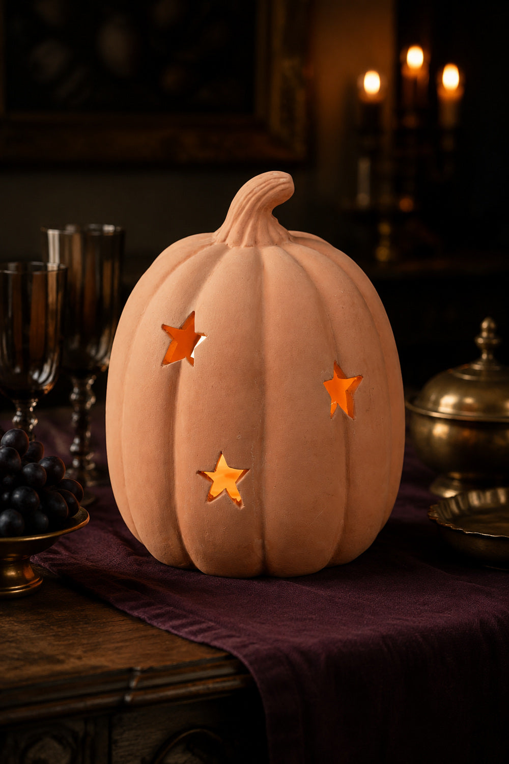 A decorative terracotta pumpkin-shaped light with three star-shaped cutouts, glowing warmly on a dark tablecloth with candles and dining props in the background.