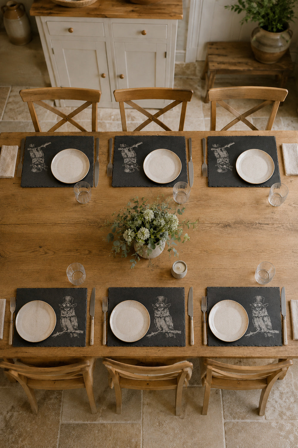 An overhead view of a rustic wooden dining table set for six, featuring dark slate placemats with a white graphic print, white plates, and wooden chairs in a warm, neutral-toned dining room.