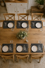An overhead view of a rustic wooden dining table set for six, featuring dark slate placemats with a white graphic print, white plates, and wooden chairs in a warm, neutral-toned dining room.