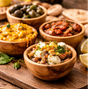 Assorted tapas in wooden bowls on a wooden surface with olives and bread in the background.
