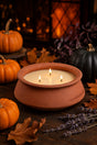 A terracotta clay candle holder with three lit candles, styled on a dark wooden table with autumn leaves, small pumpkins, and lavender sprigs.