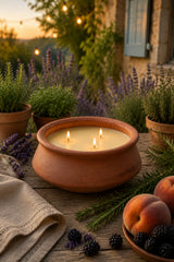 A terracotta clay candle holder with three lit wicks filled with creamy yellow wax, set on a rustic wooden table surrounded by lavender, blackberries, peaches, and greenery in an outdoor garden setting at sunset.