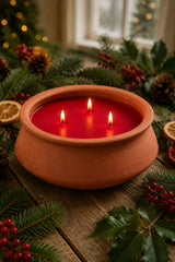 A terracotta-style bowl candle holder with three lit wicks filled with bright red wax, set on a rustic wooden surface surrounded by pine branches, holly leaves, red berries, and dried orange slices in a cozy holiday setting.