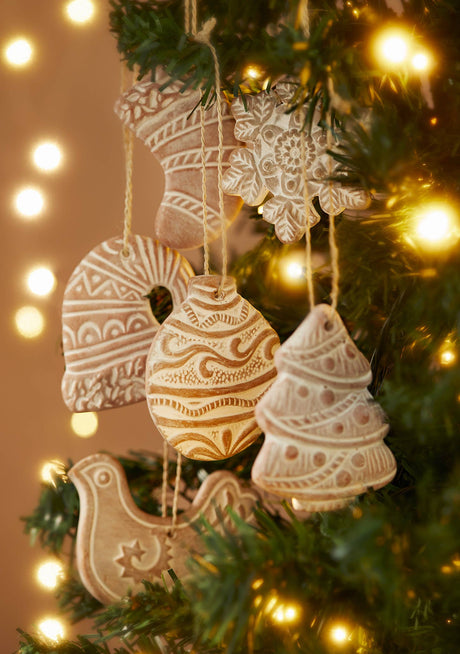 A set of hand-painted terracotta Christmas ornaments hanging on a decorated Christmas tree with warm string lights. The ornaments include a stocking, snowflake, angel, bird, and tree shapes in a light cream glaze with brown painted patterns.