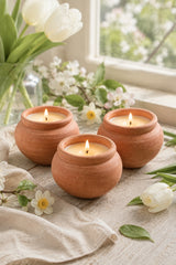 Three small terracotta clay candle holders with lit candles, arranged on a light fabric surface near a window with white tulips and blossoms.