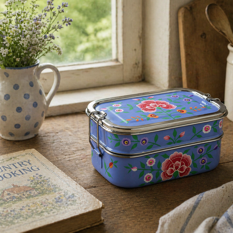 Floral-patterned metal lunch box on a wooden table with a window and kitchen items in the background.