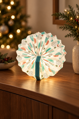 Turquoise Geometric Velvet Accordion Lamp glowing on a walnut console with Christmas tree lights softly blurred behind.