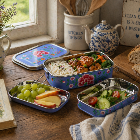 Three metal lunch boxes with food on a wooden table with a teapot and flowers in the background.