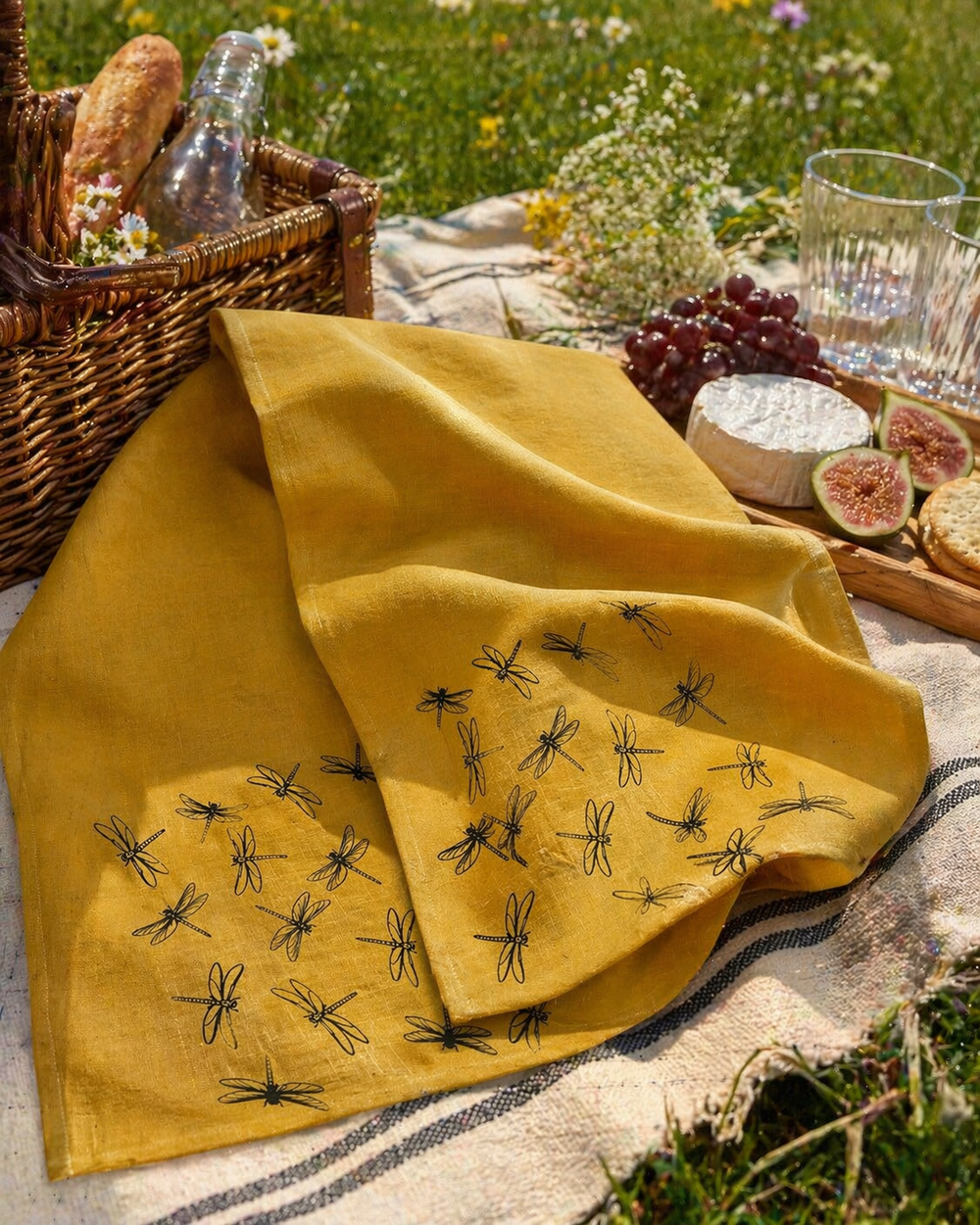 yellow Linen tea towel with black dragonfly patterns on a picnic setup with bread, cheese, and fruit.