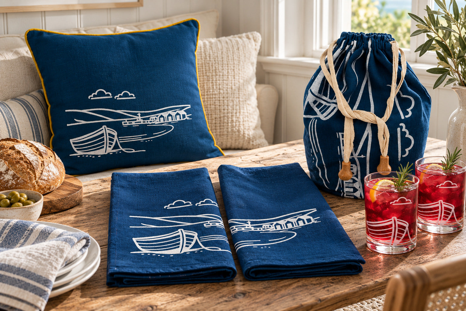 Fjære navy coastal cushion, printed tea towels, bread bag and boat etched glass tumblers arranged together.